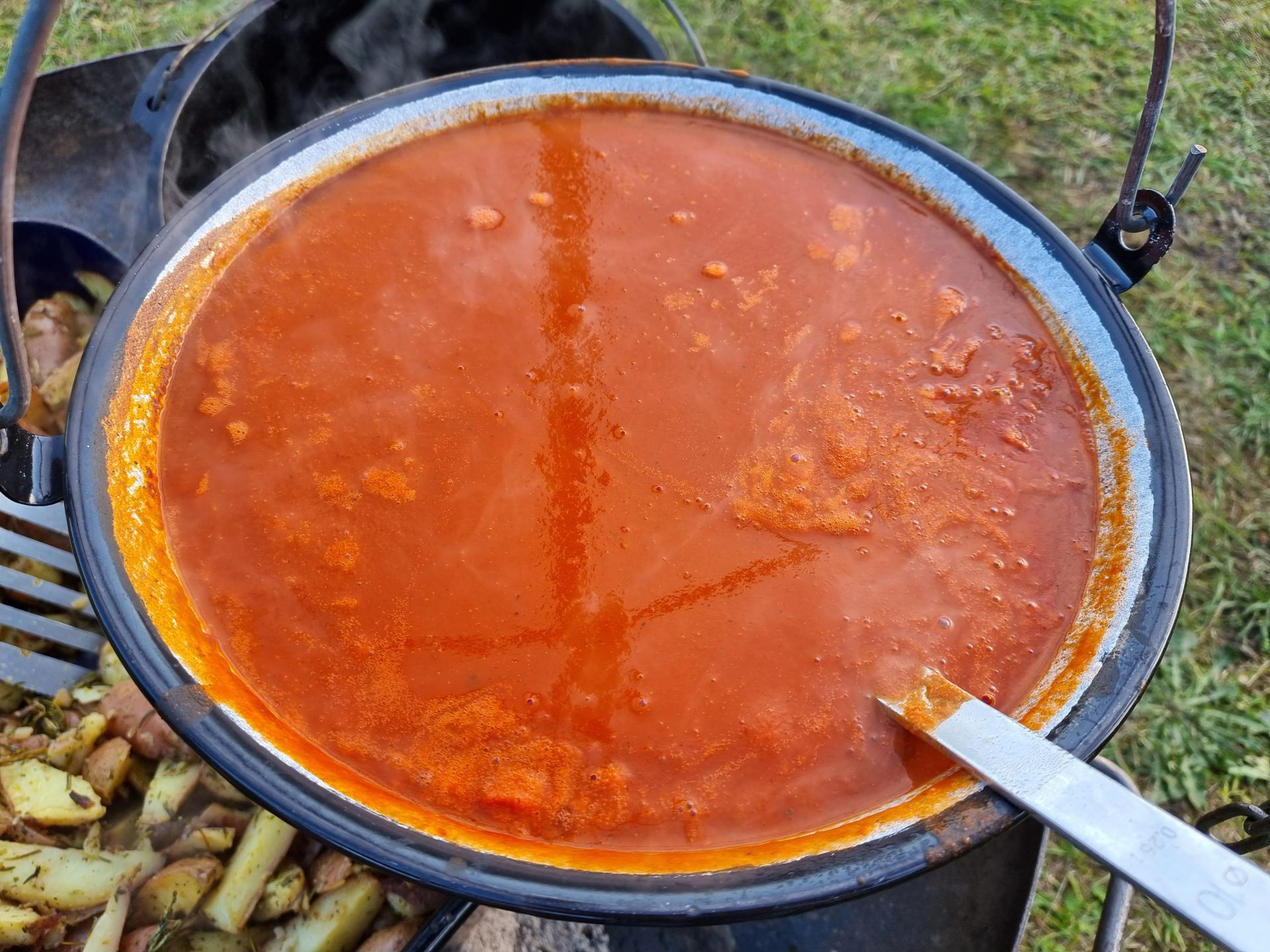 Een grote pan rode tomatensoep staat buiten te pruttelen met aardappelen eronder.