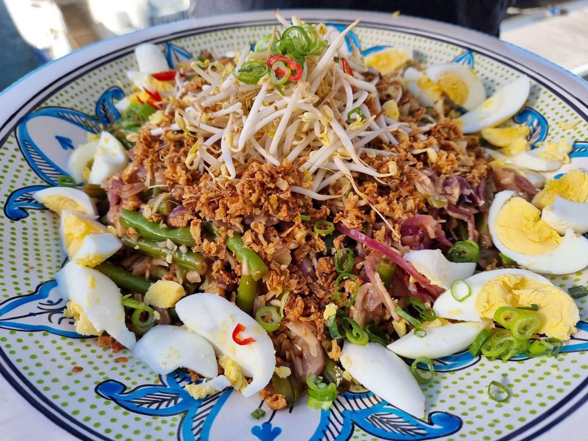 Gadogadosalade met taugé, gehakte noten, sperziebonen, gekookt ei en lente-uitjes op een bord met een decoratief patroon.