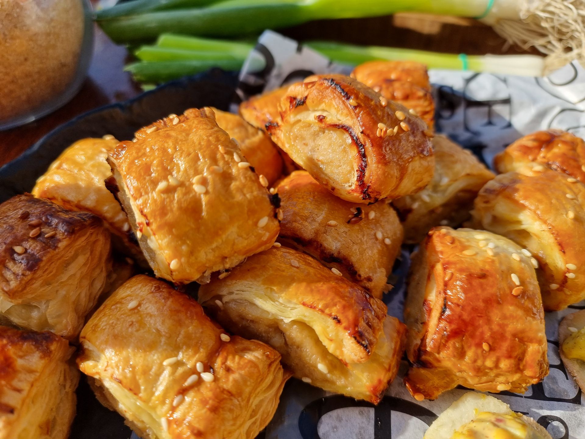 Goudbruine, bladerige mini saucijzenbroodjes met sesamzaadjes, opgestapeld in een mandje naast lente-uitjes.