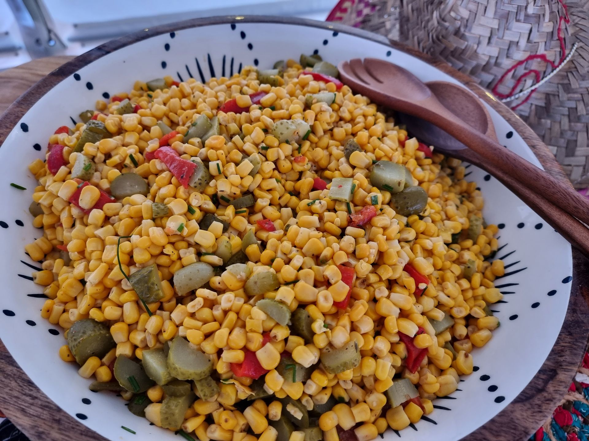 Maissalade met augurken en rode paprika in een kom met patroon, houten serveerlepels.