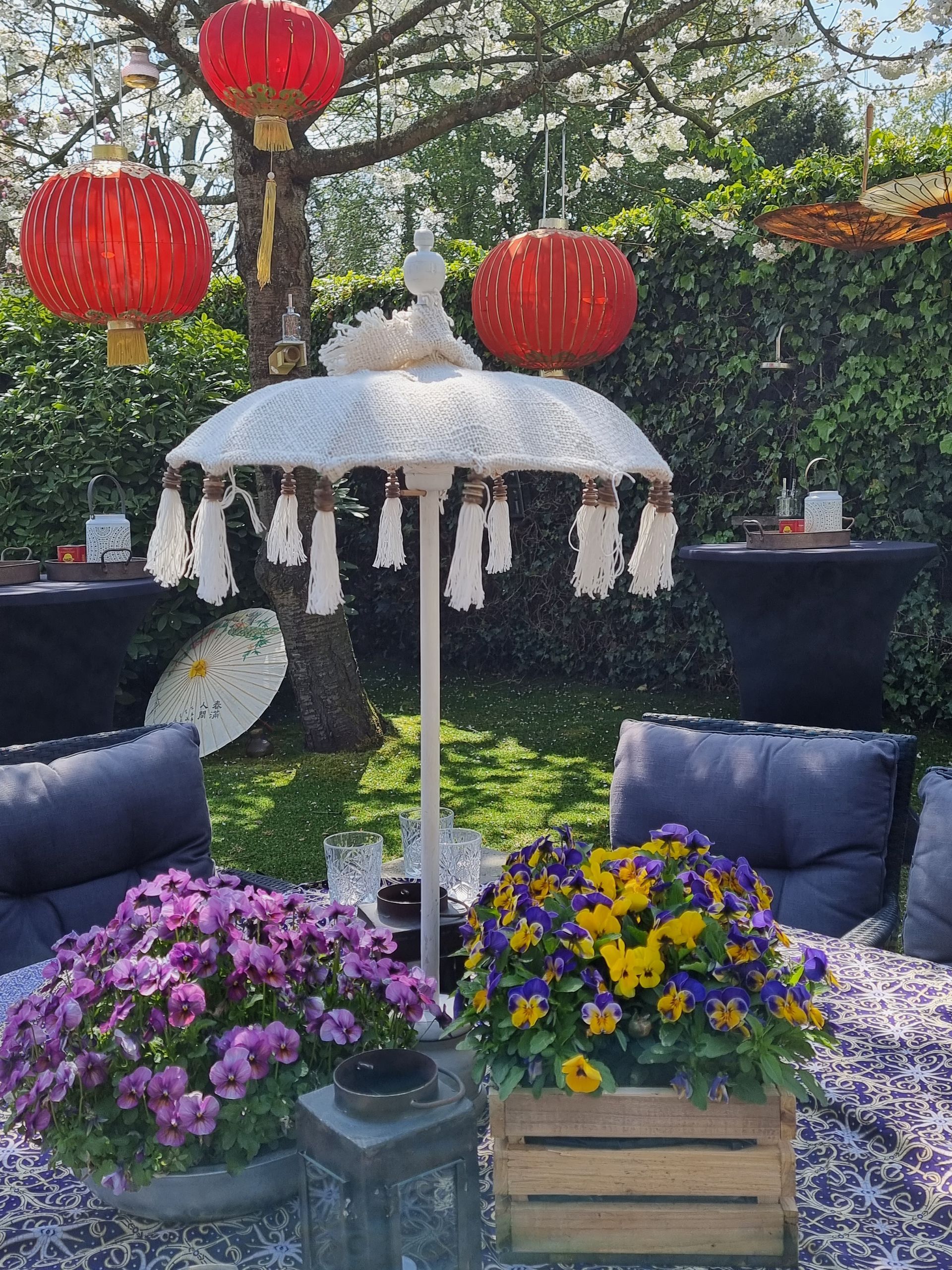 Een terras in Aziatische stijl met rode lantaarns, een decoratieve parasol, bloemen en zachte kussens.