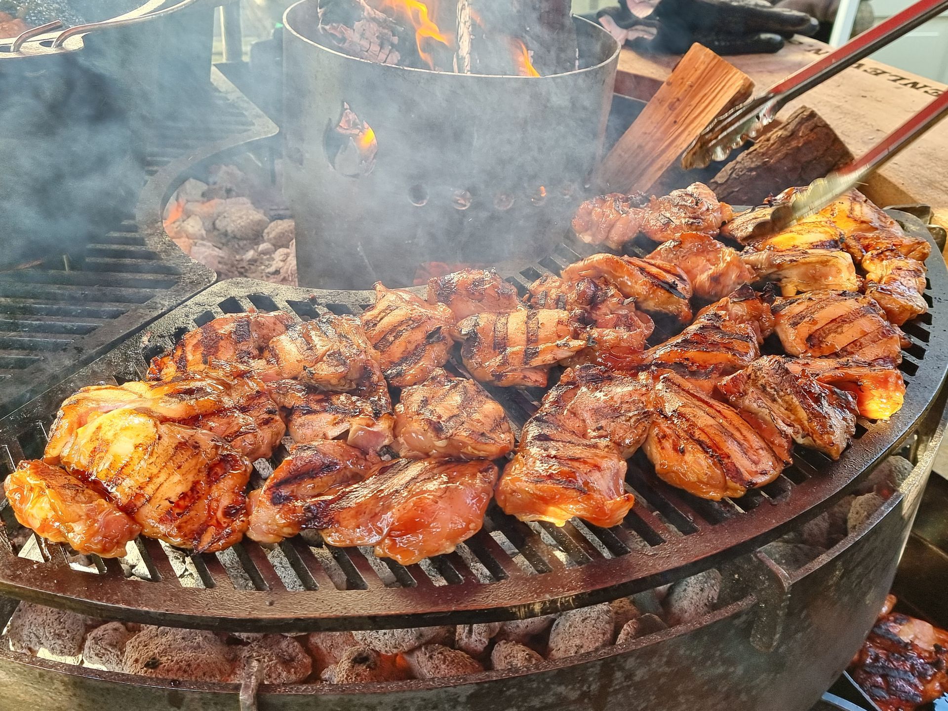 Gegrilde kipstukken op een grote ronde grill, rook die opstijgt, met een vuur zichtbaar op de achtergrond.