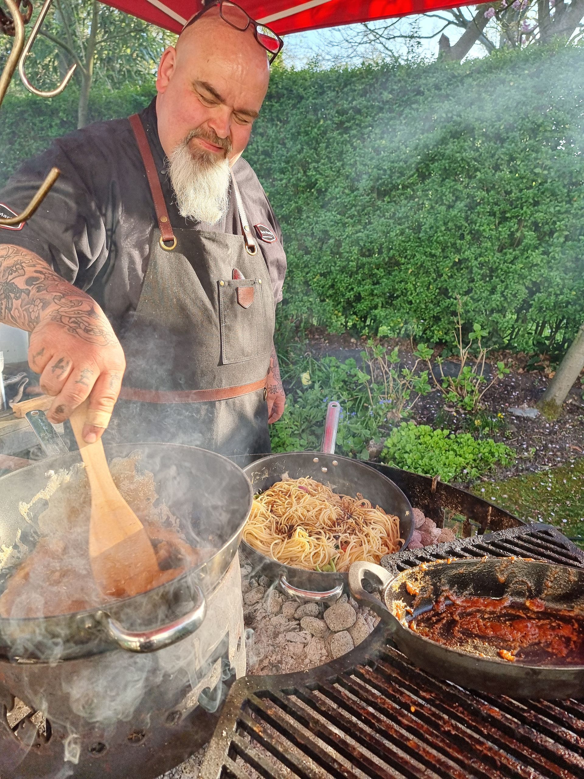 Chefkok Martin roert voedsel in een pan boven een grill in de buitenlucht. Er stijgt rook op.