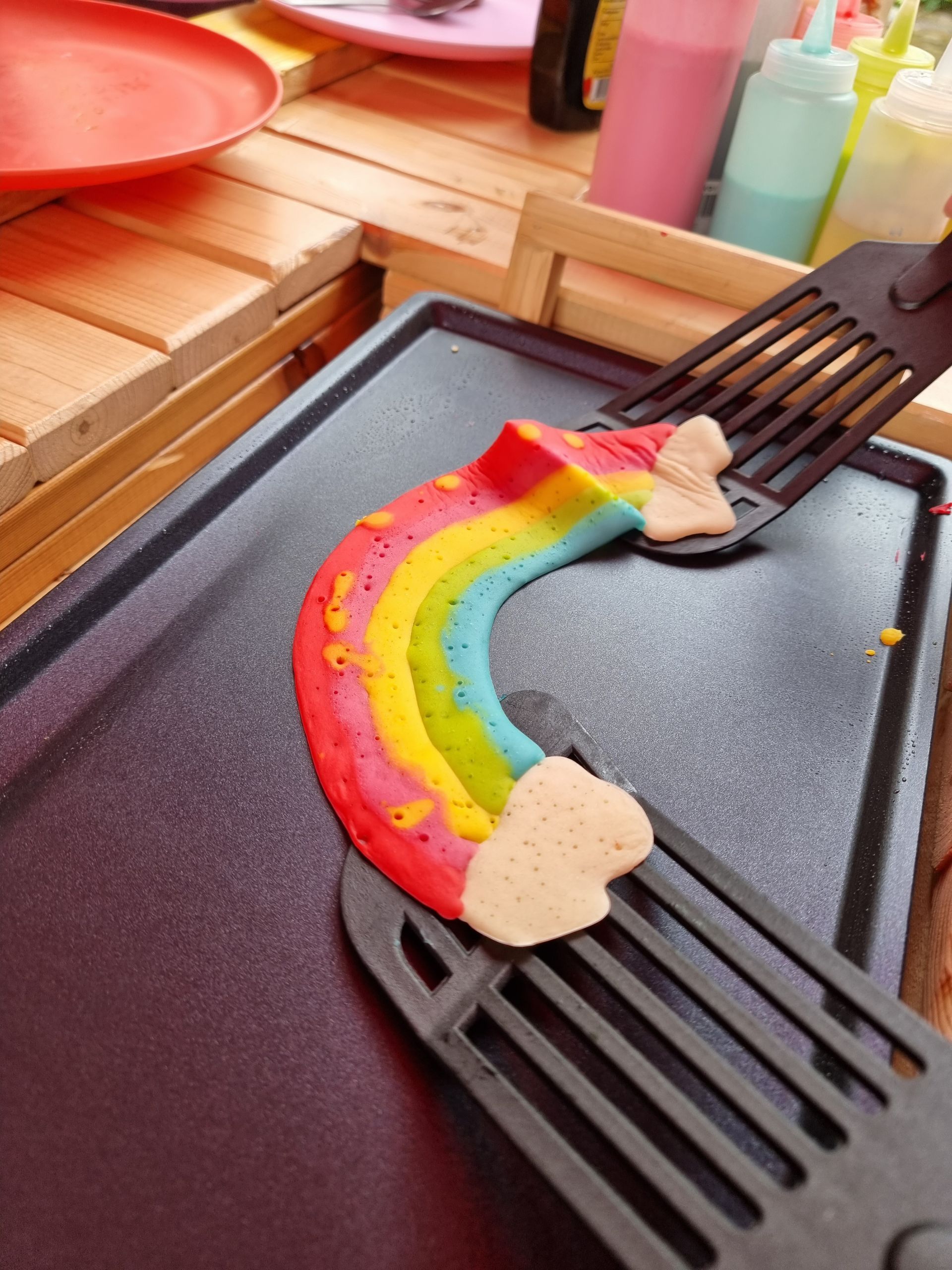 Regenboogpannenkoek op een bakplaat, vastgehouden door twee spatels. 