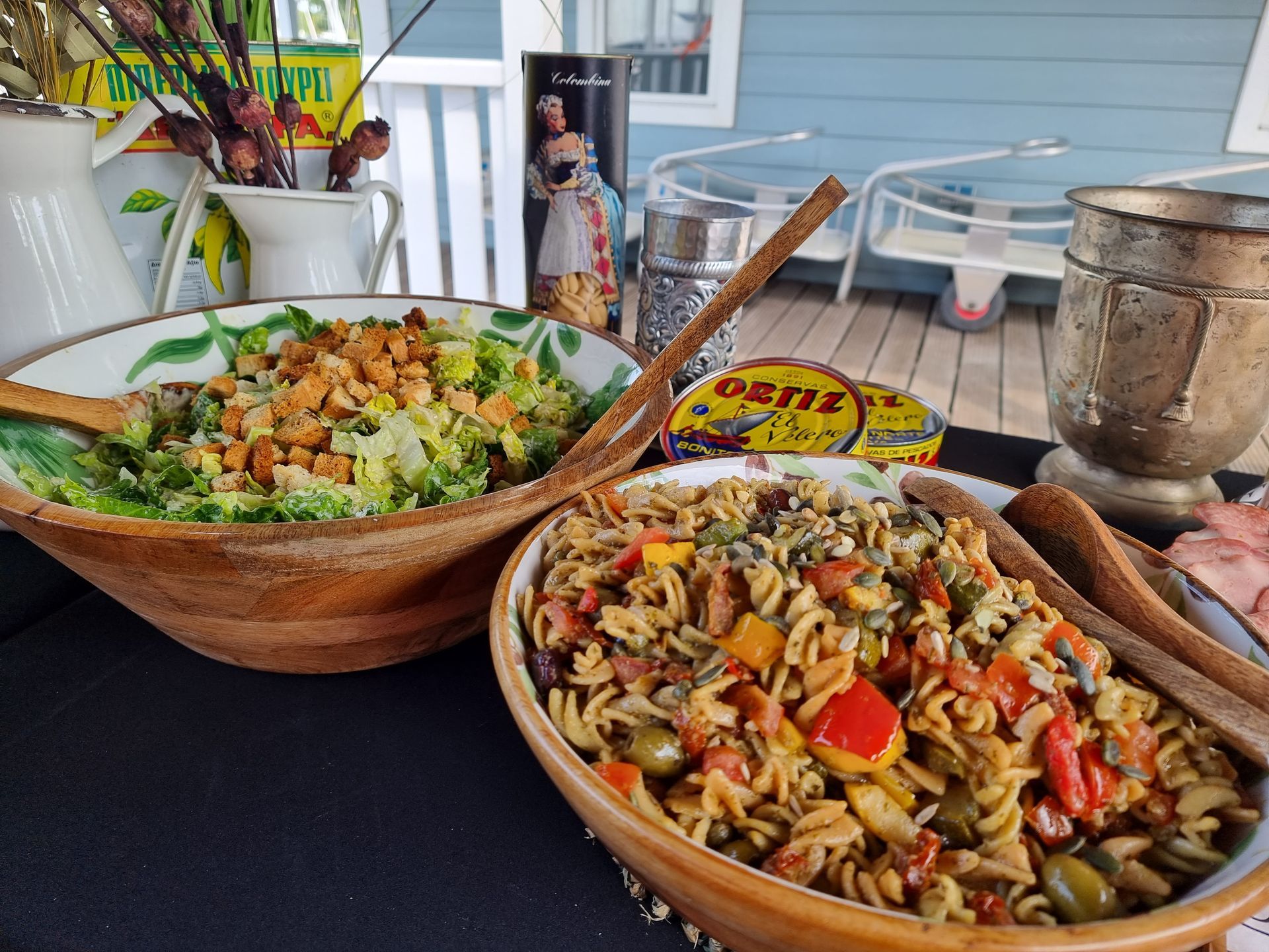 Twee houten kommen met eten: salade met croutons en pasta met groenten, op een terras.