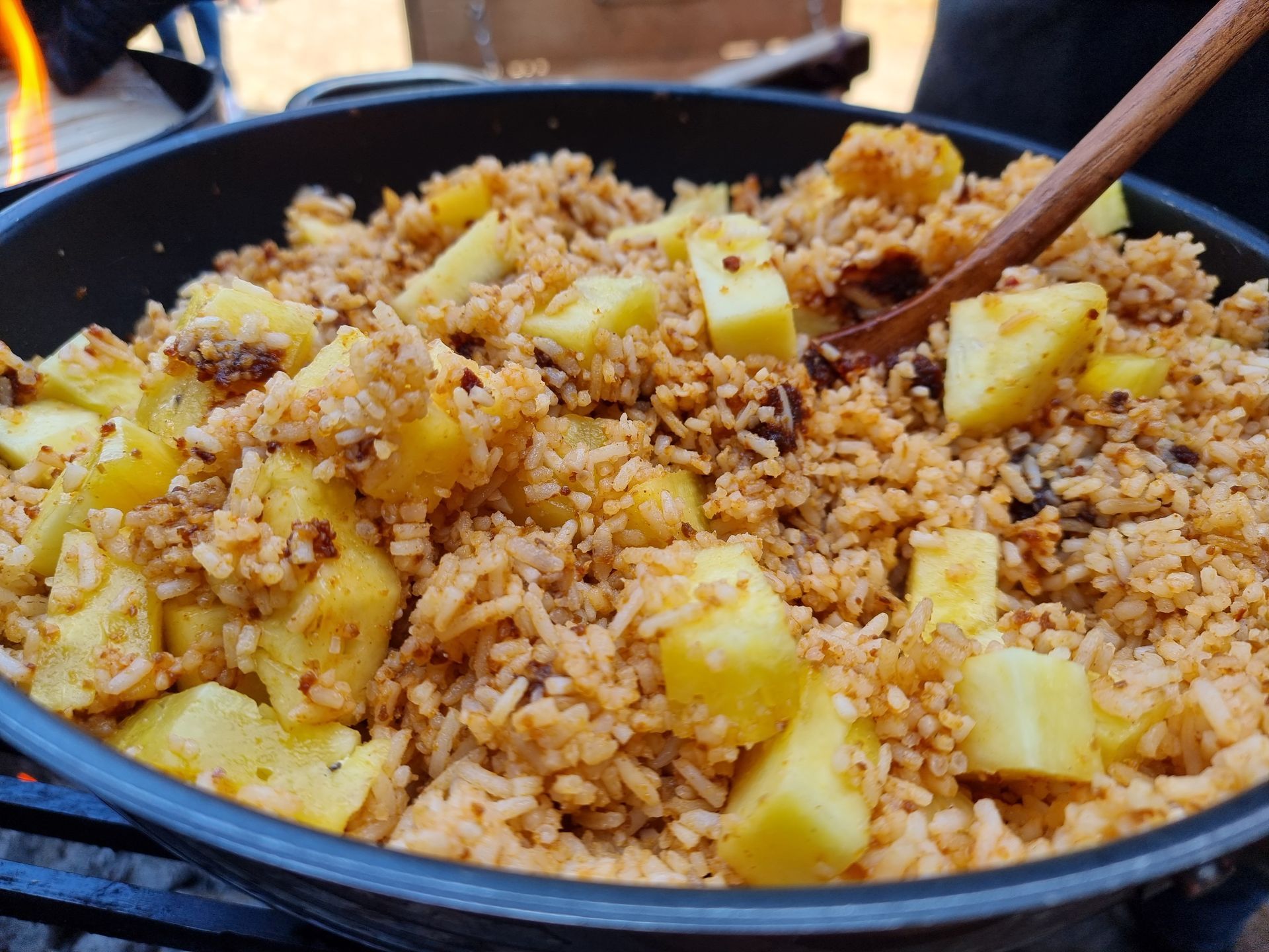 Rijstgerecht met ananasstukjes, bereid in een gietijzeren pan boven een vuur, met een houten lepel in het gerecht.