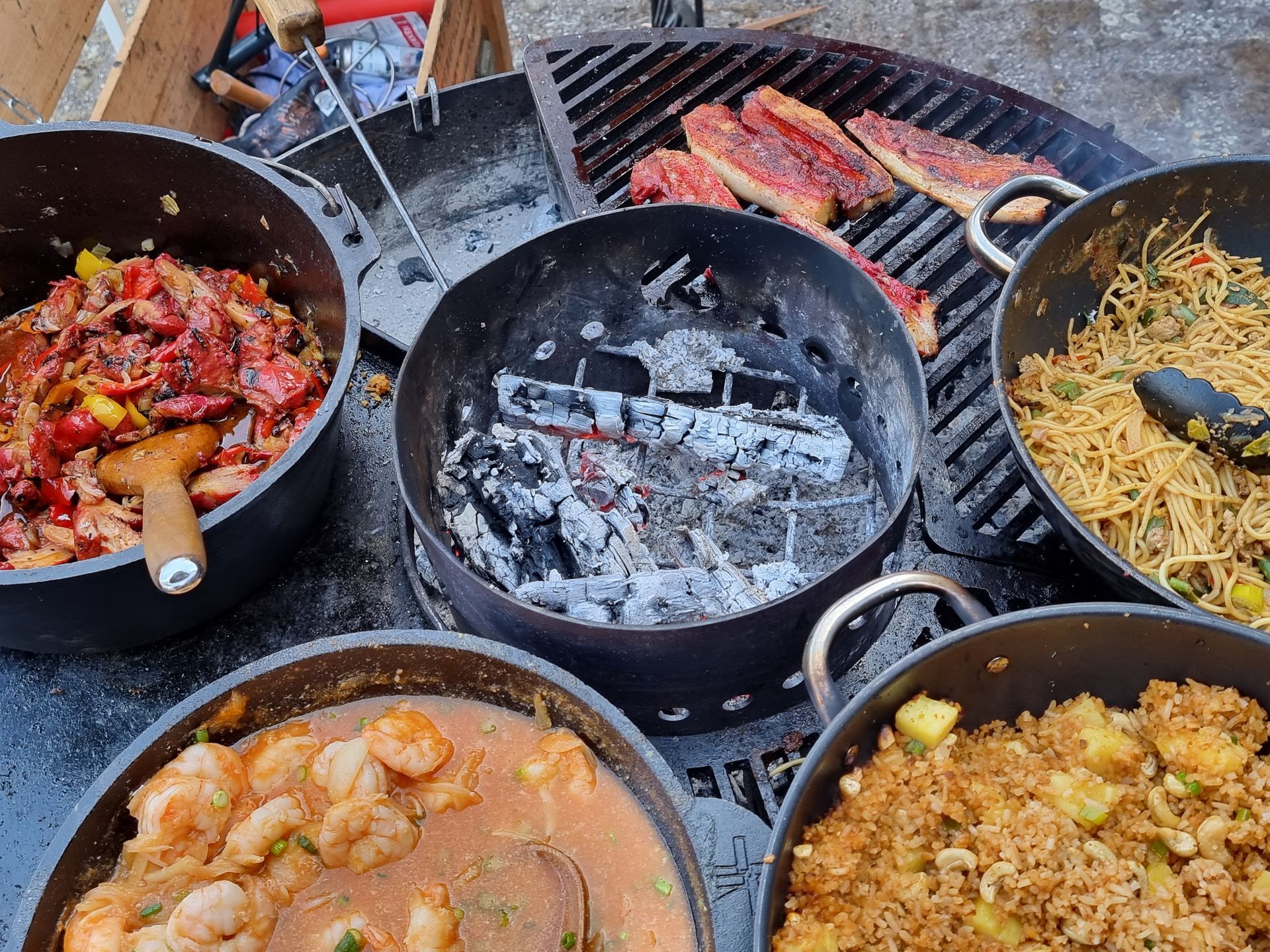 Buitenkeuken met grill en diverse zwarte potten met bereid voedsel, waaronder vlees, garnalen en noedels.