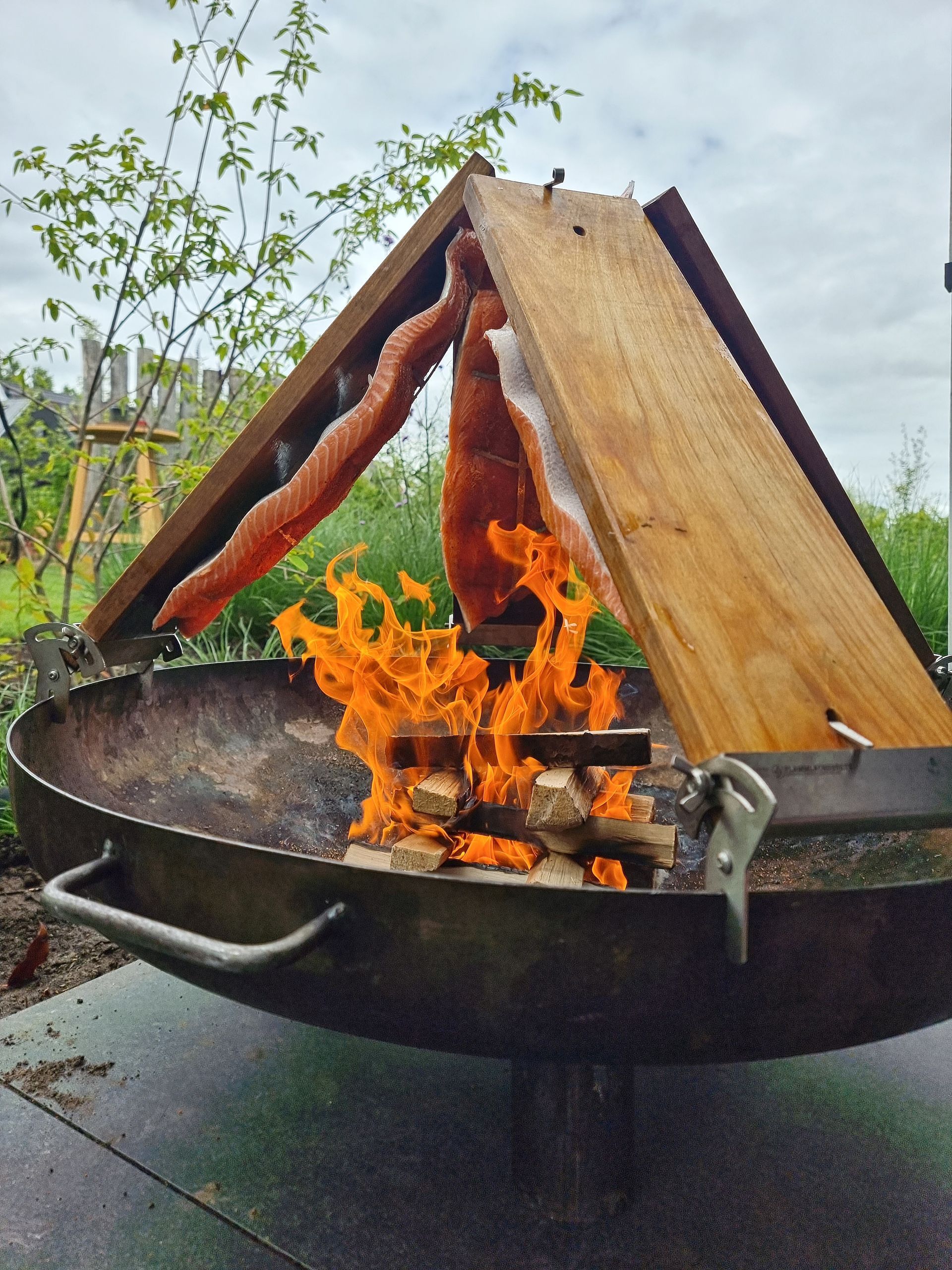 Zalm die boven een open vuur in een metalen vuurschaal wordt gegaard. 