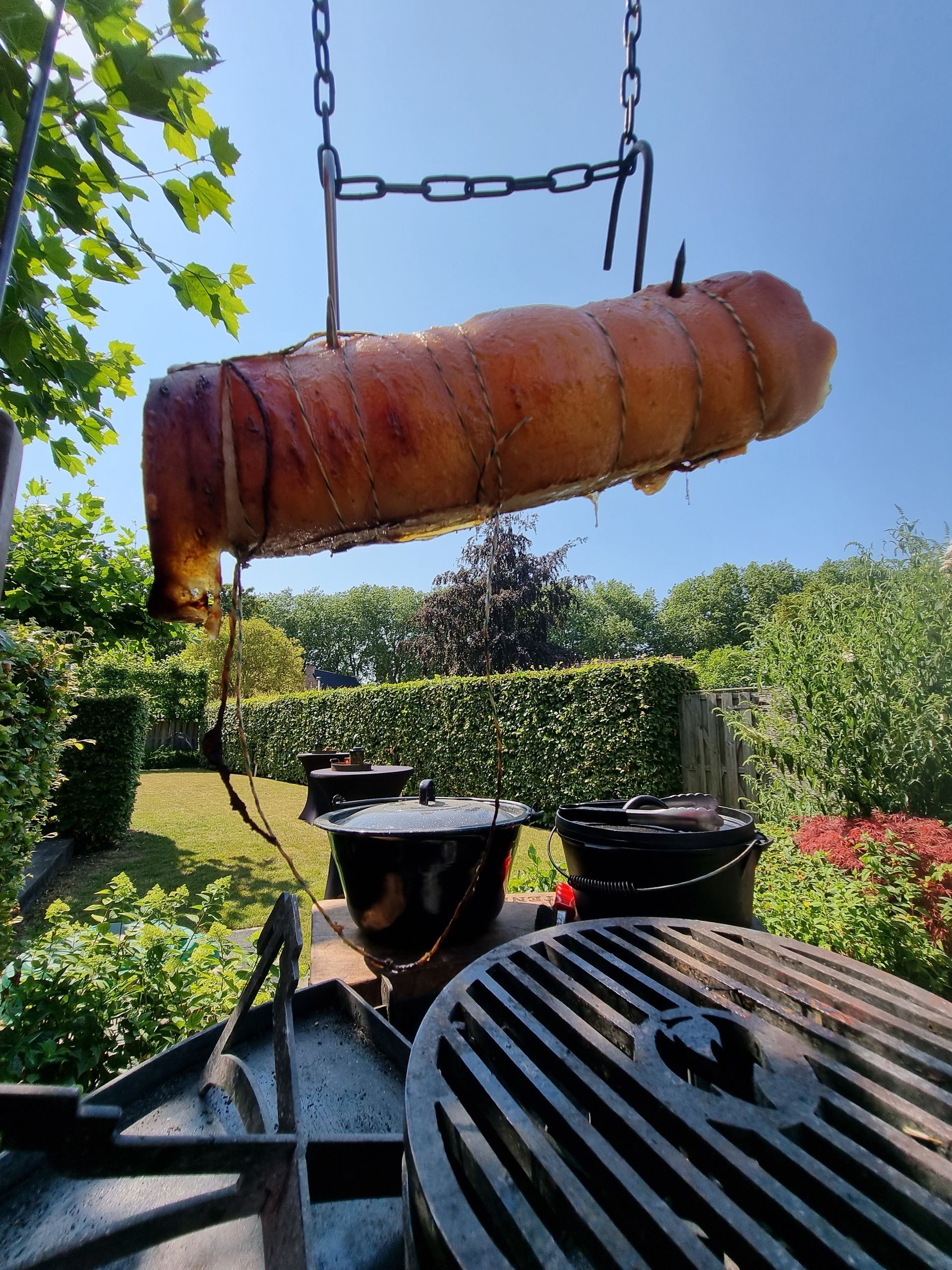 Groot braadstuk, hangend boven een grill, buiten. Zonnige dag, groen op de achtergrond.