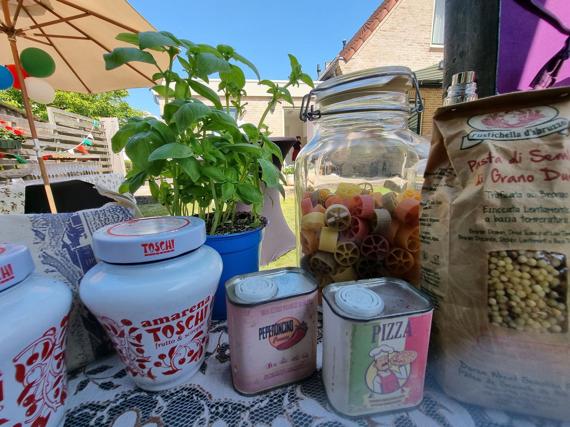 Pasta en Italiaanse etenswaren op een tafel buiten: een pot pasta, een basilicumplantje, blikjes en een zak pasta.