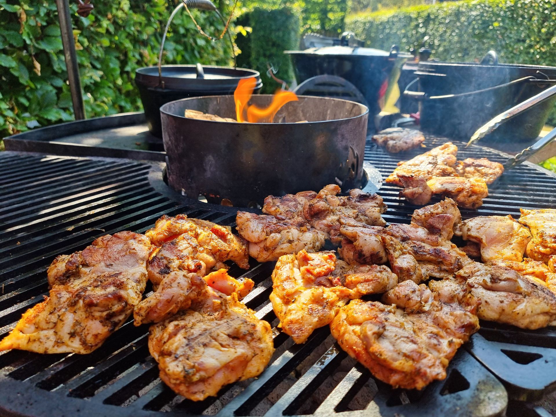 Gegrilde kip die buiten op een barbecue met vlammen wordt bereid, omringd door braadpannen.