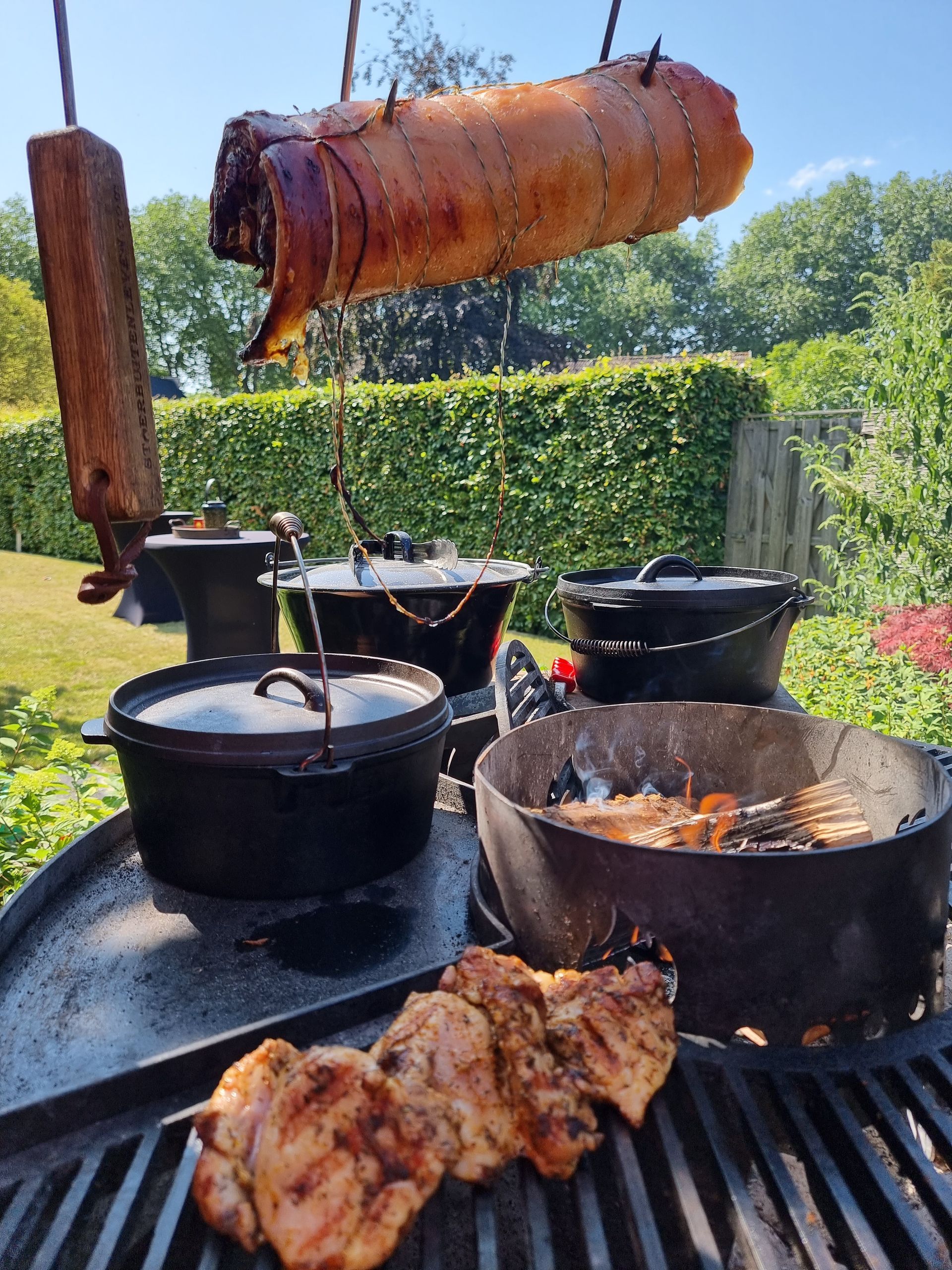 Buiten koken: gebraden vlees boven gietijzeren pannen en gegrilde kip. Zonnige dag, groen gebladerte.