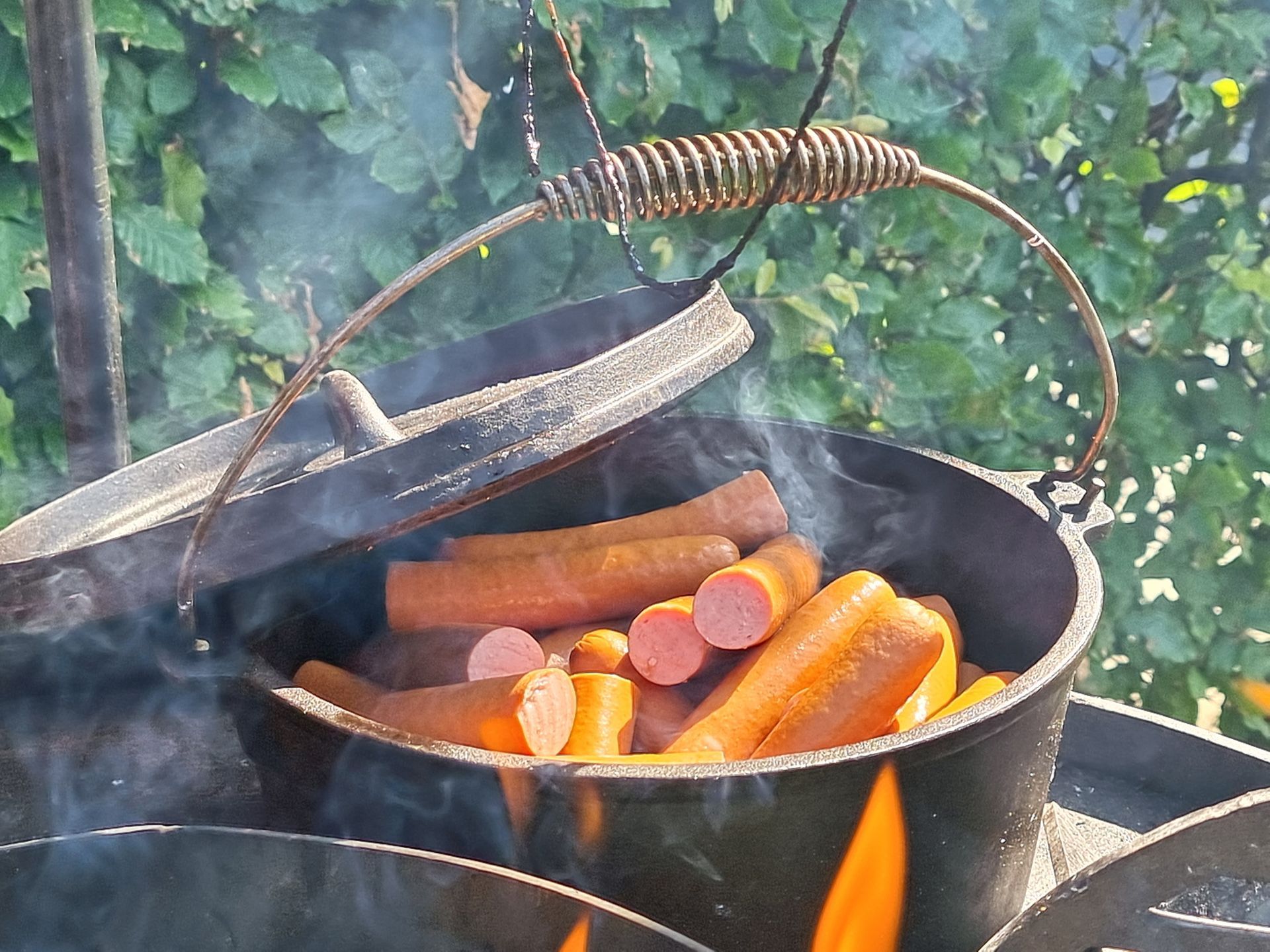 Hotdogs die in een braadpan boven een open vuur worden verwarmd en stoom afgeven tegen een groene achtergrond.