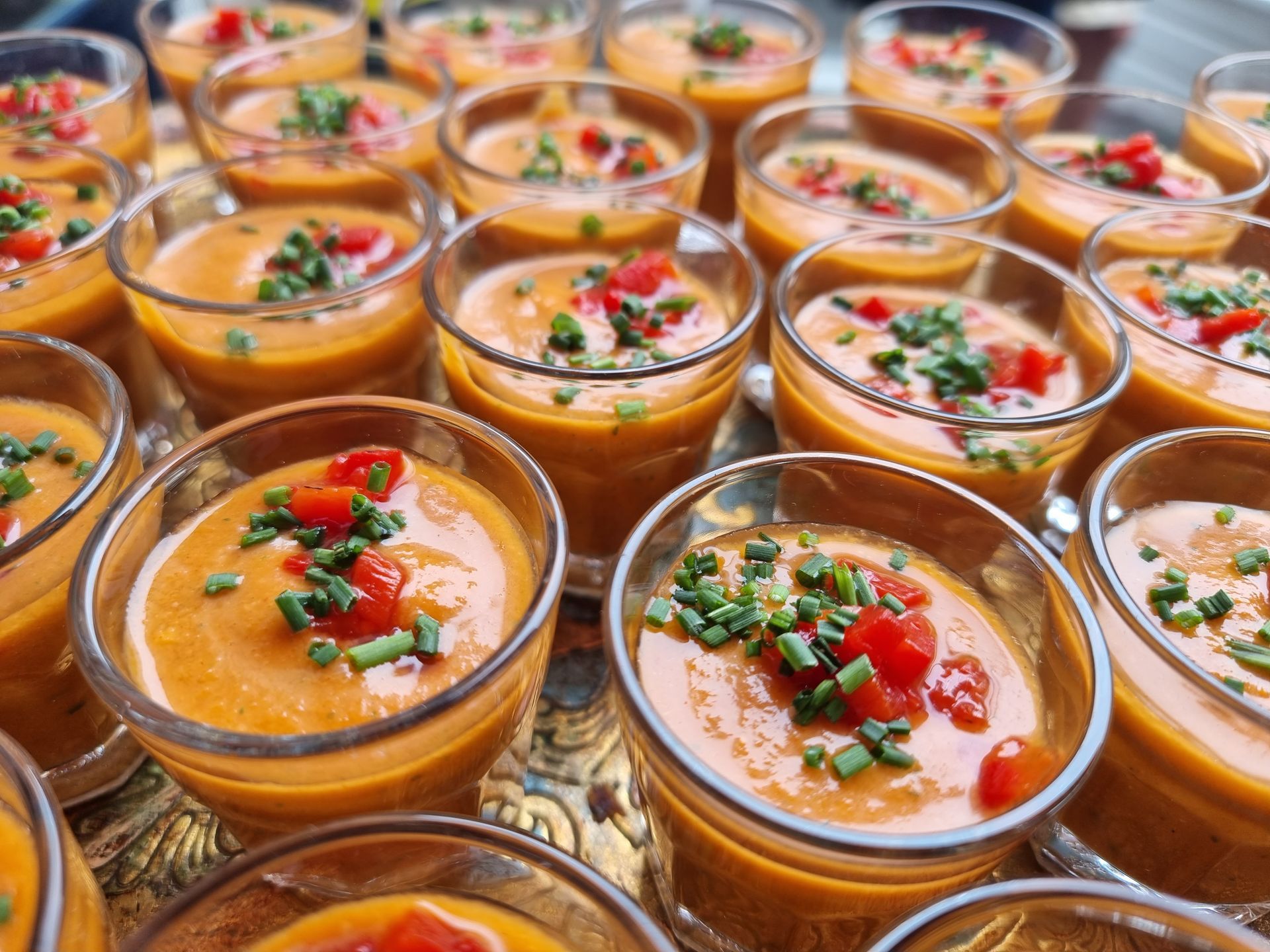 Kleine glaasjes soep, gegarneerd met rode paprika en groene bieslook, op een dienblad.