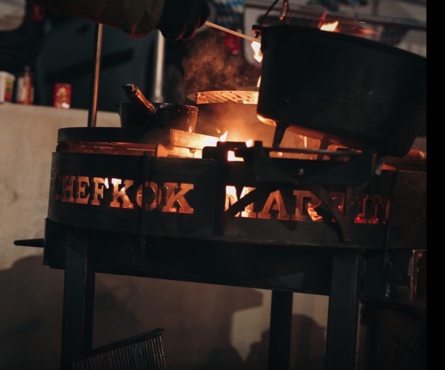 Chefkok Martin kookt boven een open vuur, gebruikmakend van een op maat gemaakte vuurkorf met de tekst 