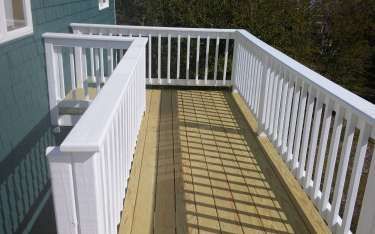 White-railed wooden deck with vertical balusters. Light brown wood flooring. Green building wall on the left.