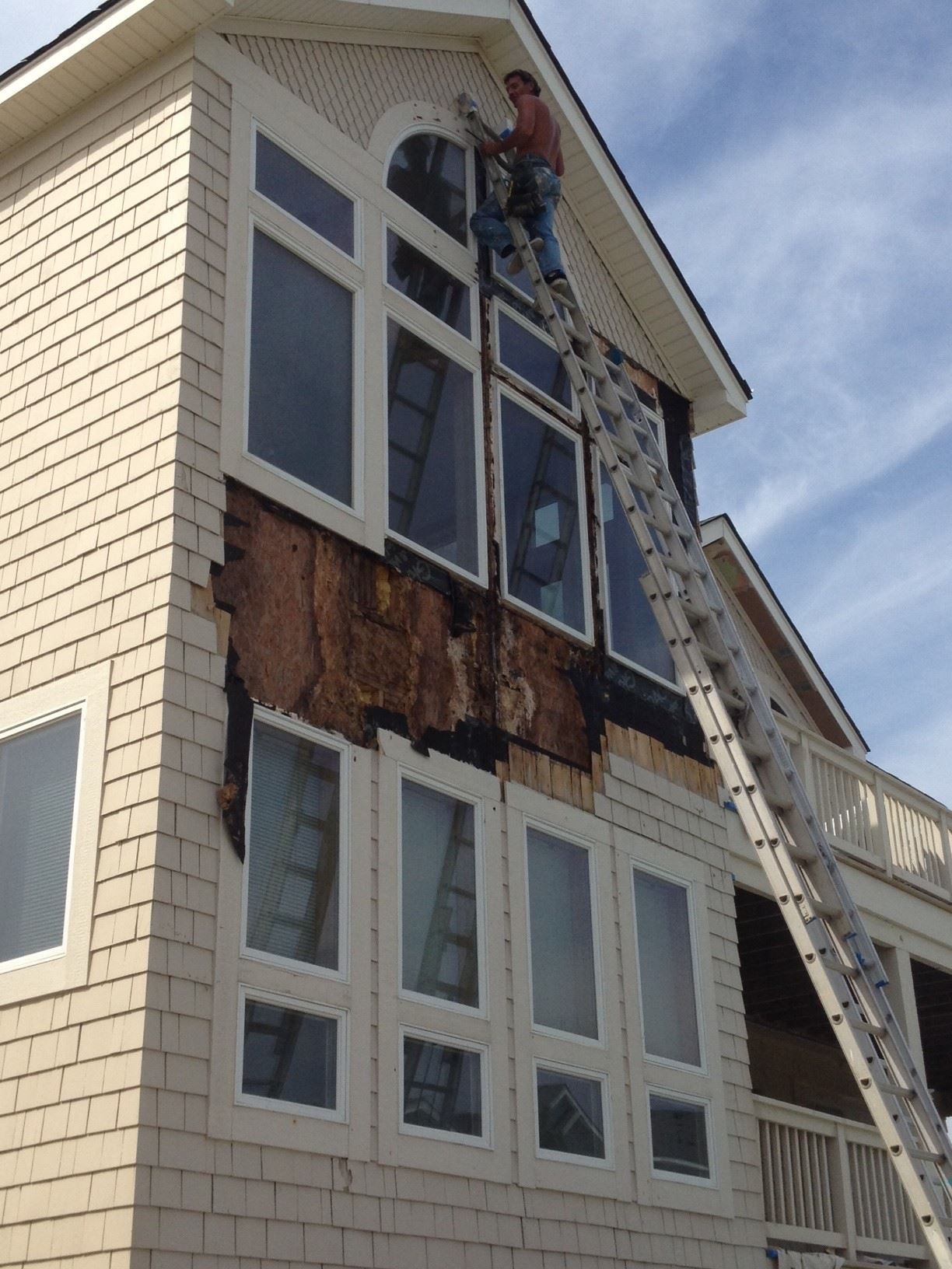 Person on ladder repairs house siding with damaged section. Tall windows, blue sky.