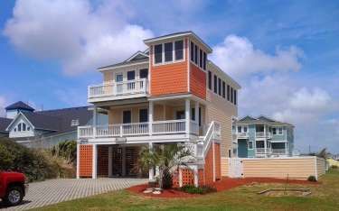 Coastal home with orange and tan accents, balconies, and elevated foundation on a sunny day.