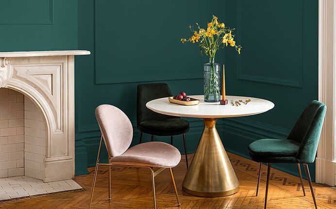 Dining room with teal walls, marble-topped table, gold base, velvet chairs, and flowers.
