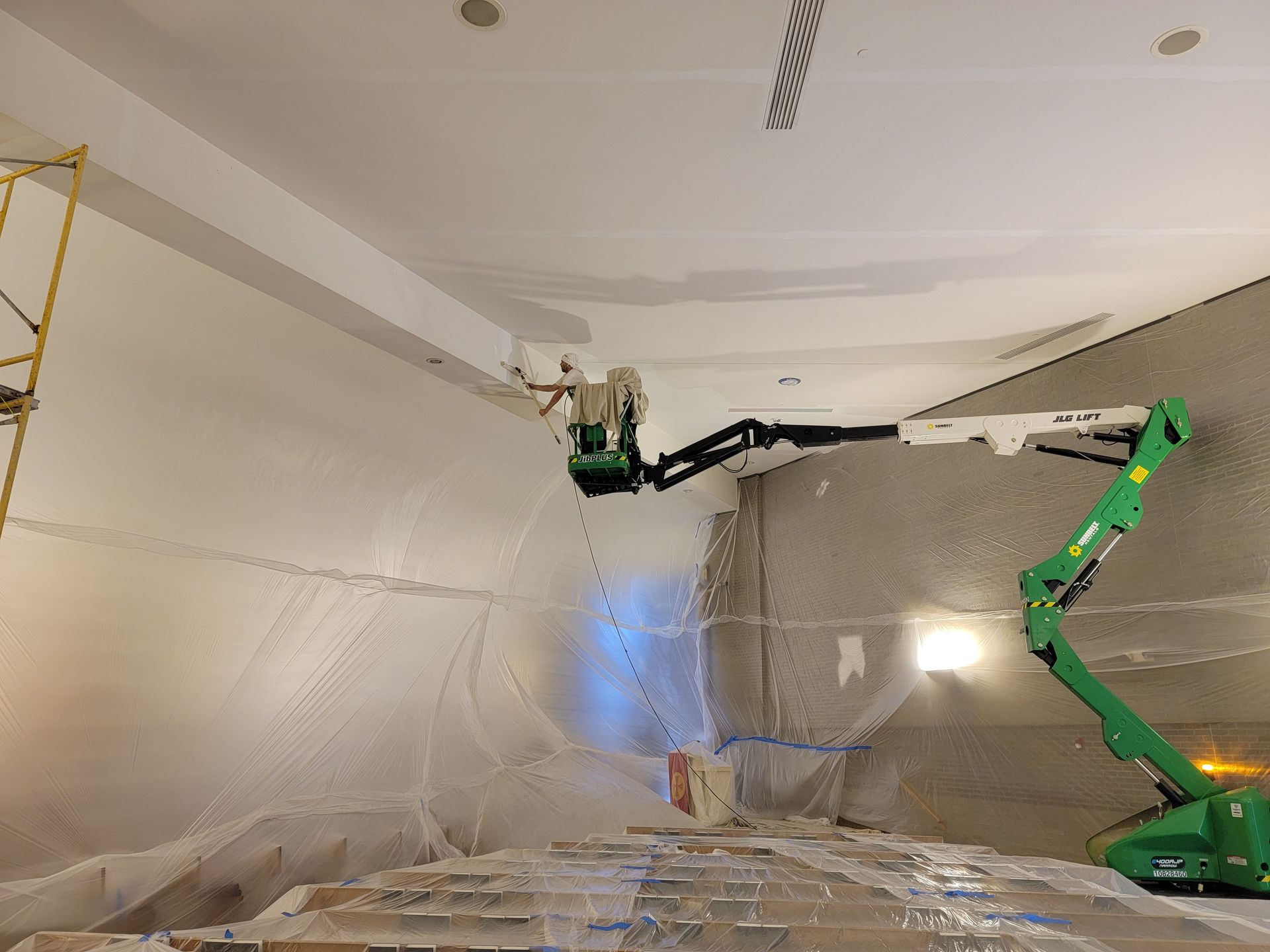 Person painting a high ceiling from a green boom lift in a construction site.