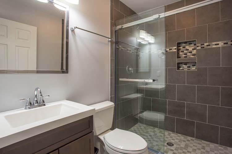 Modern bathroom with gray tiled walls, glass shower, white sink, and toilet.