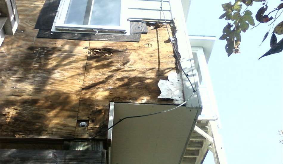Damaged exterior building corner with exposed wood and siding, near a window.