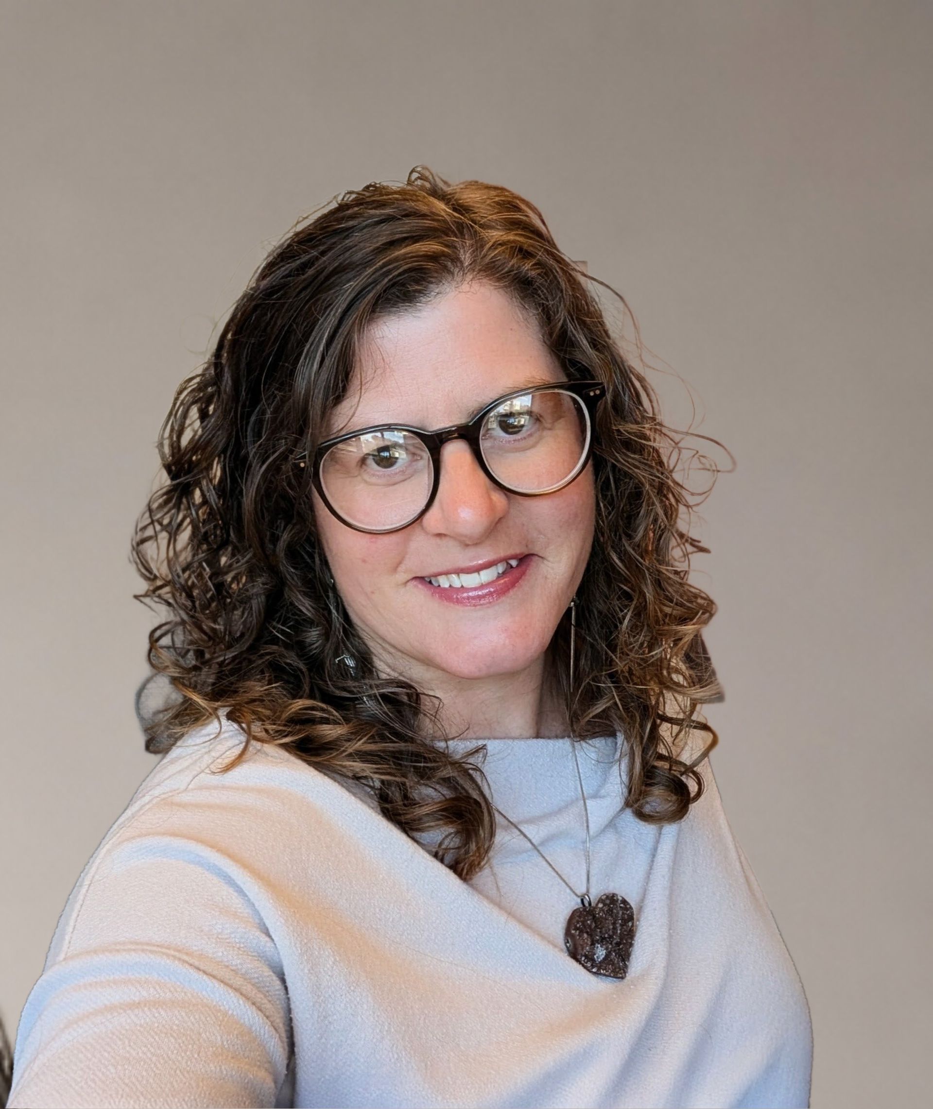 Woman with curly hair and glasses smiles. Interior, cross on wall, shelves with pictures visible.