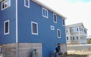 Blue house with white trim and multiple windows, adjacent to a beige house, both near a beach.