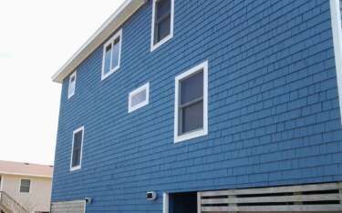 Blue-sided two-story house with white-framed windows, against a bright sky.