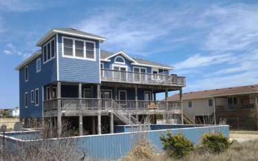Blue beach house with wooden decks under a partly cloudy sky.