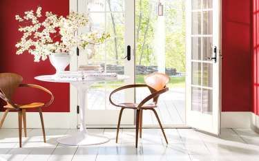 Room with red walls, white table, wooden chairs, open French doors, and a vase of white flowers.