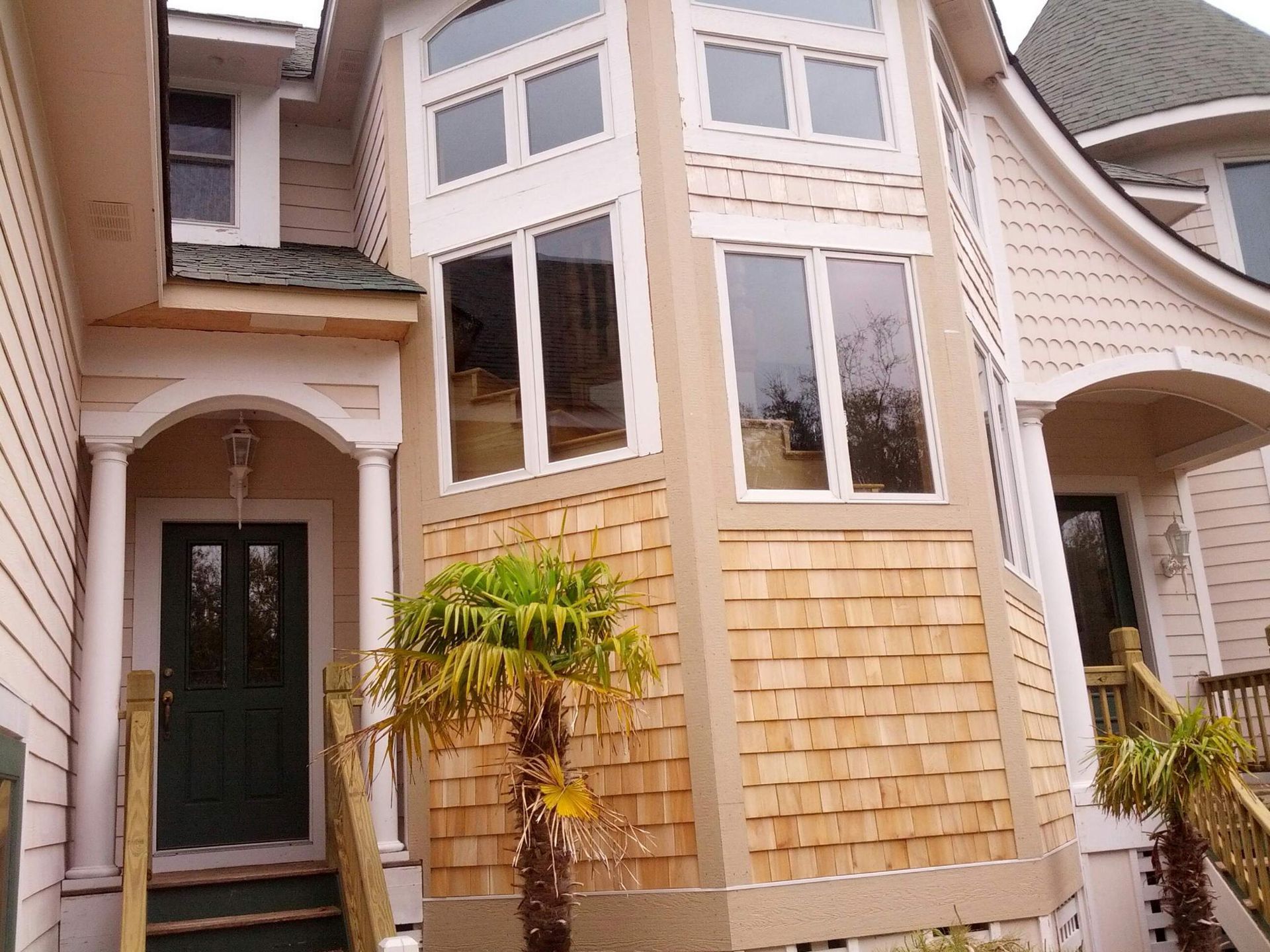 Two-story house with light brown wood shingles, many windows, and a green front door. Palm tree in front.