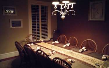 Formal dining room with long table set for a meal. Wooden chairs, chandelier, shuttered window, and art on the walls.