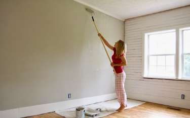 Woman painting a wall gray with a roller on a long handle in a room.