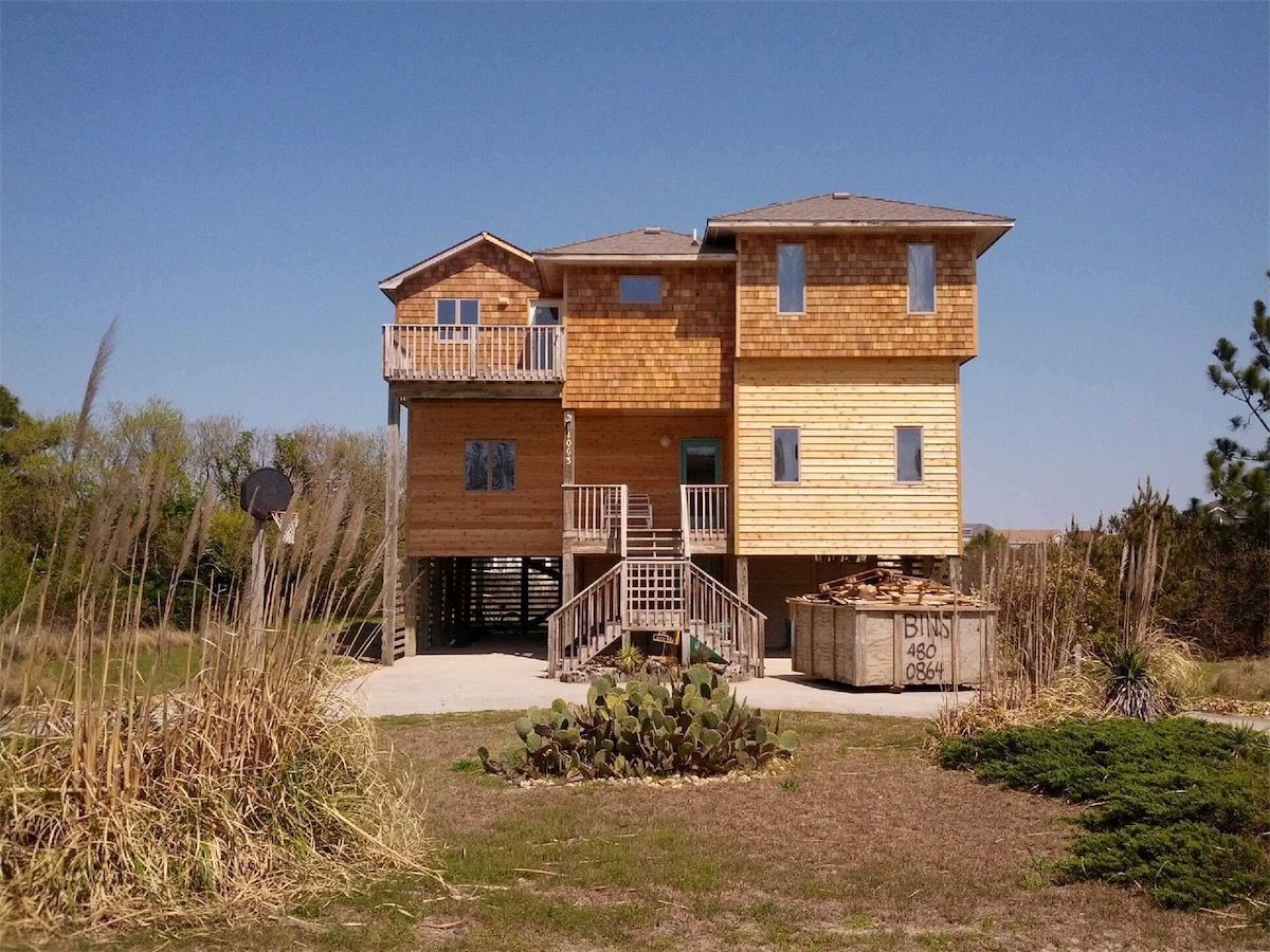 Three-story house on stilts with wooden siding and a deck, in a grassy area under a blue sky.