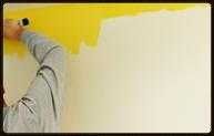 Person painting a wall yellow with a roller; a portion of the wall remains unpainted in off-white.