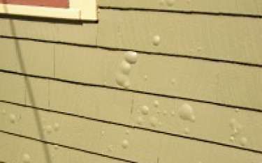 Bubbling paint on the side of a beige wooden house with a red-trimmed window.