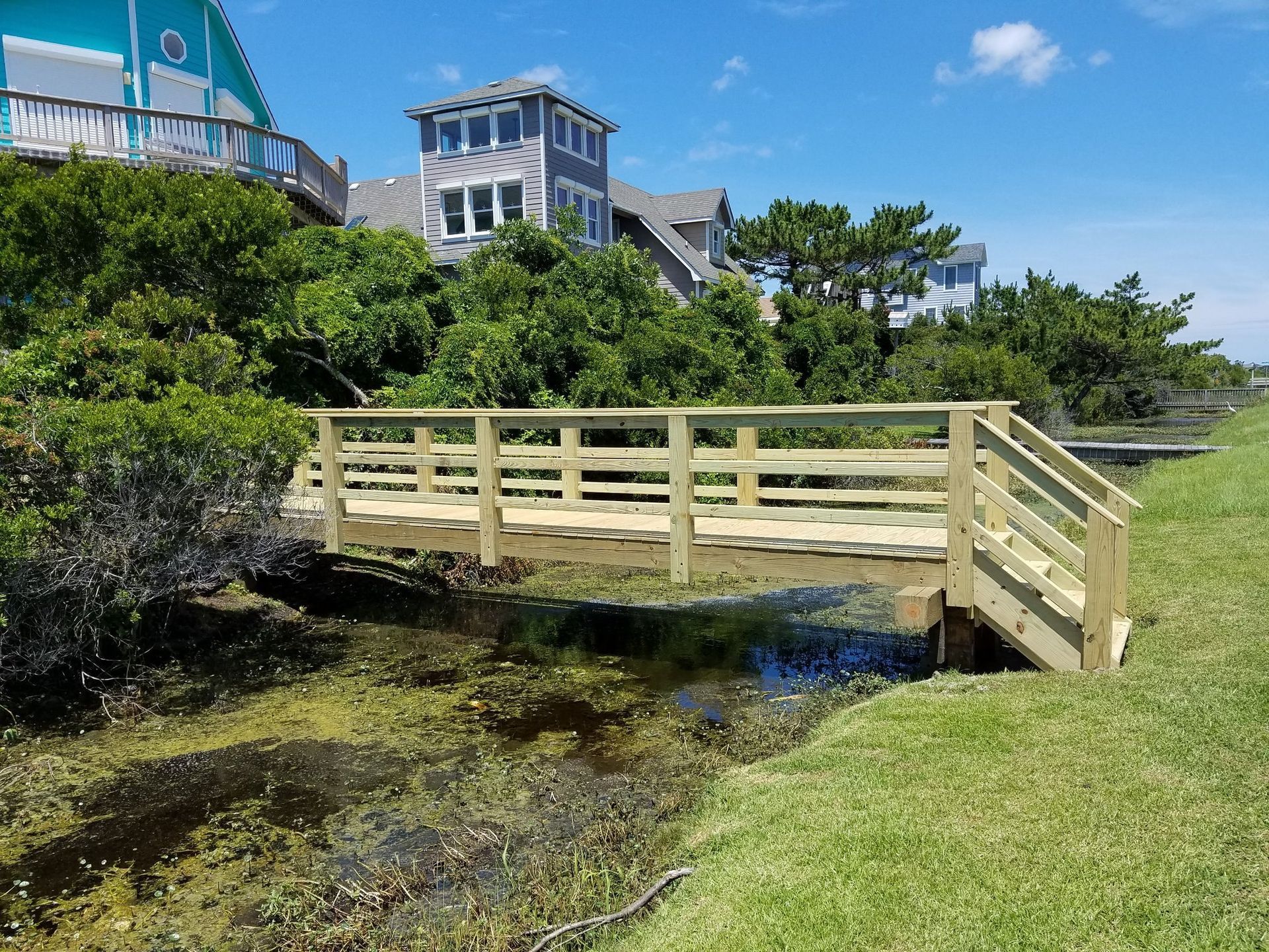 Wooden bridge over a marshy creek with houses and greenery in the background, sunny day.