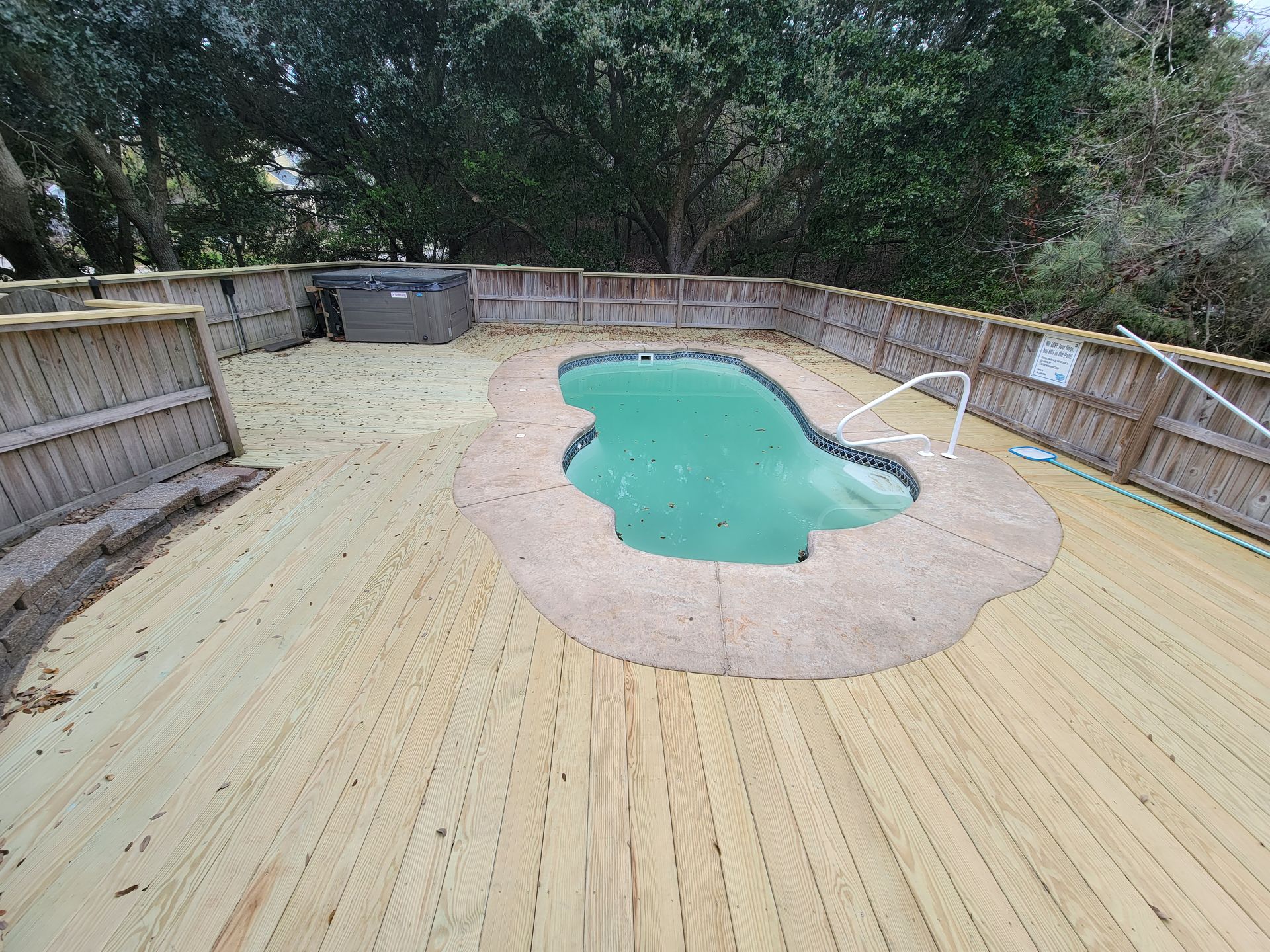 Wooden deck surrounds an irregularly shaped pool. A hot tub sits in the corner, and a fence borders the deck.