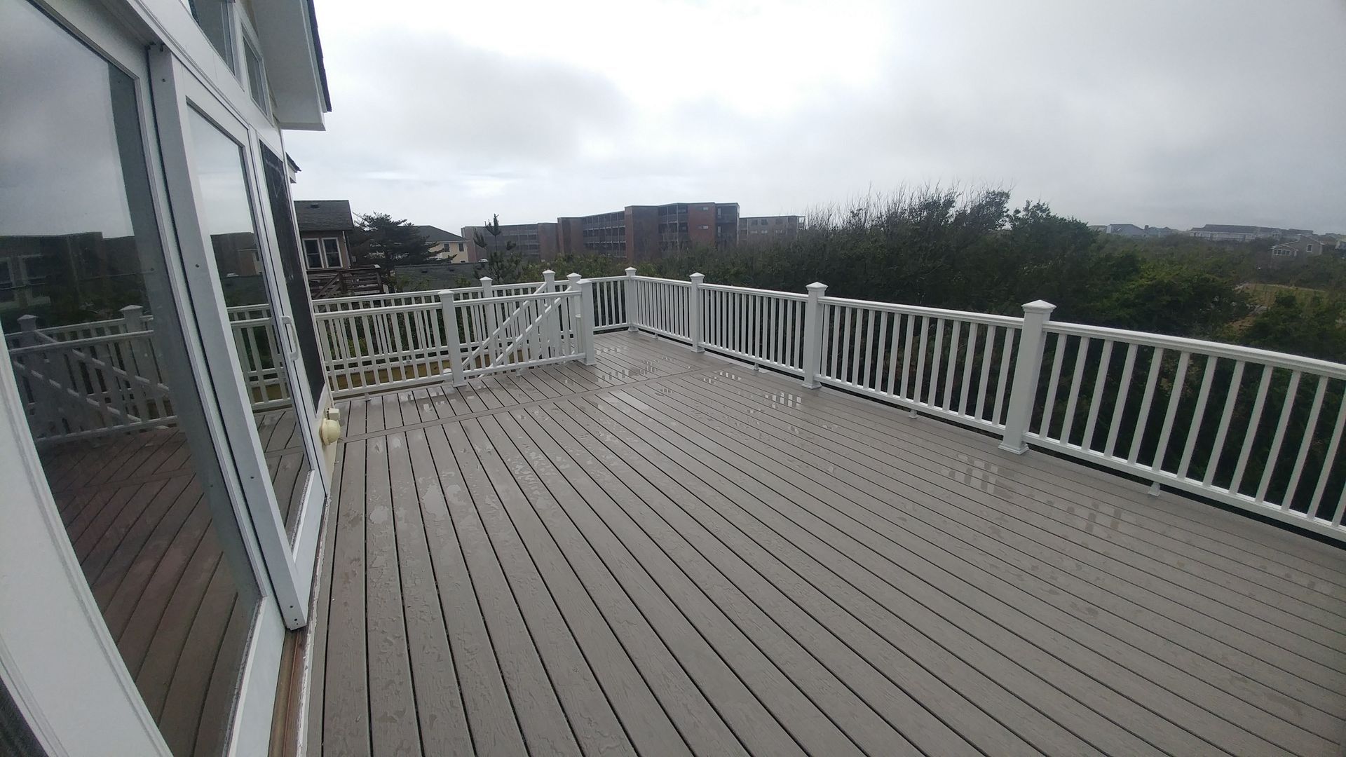 Gray deck with white railing next to glass doors, overlooking trees and buildings on a cloudy day.