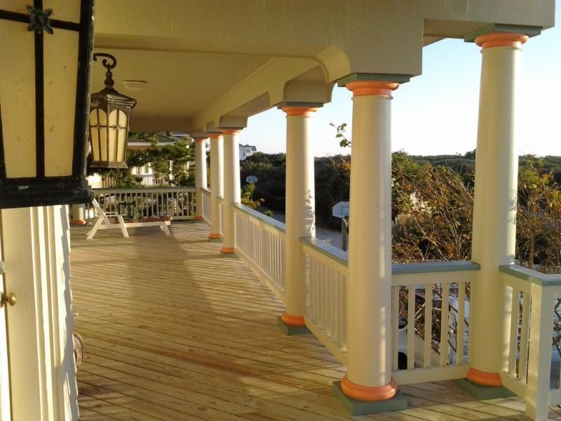 Wooden veranda with white columns and orange accents, overlooking trees and a glimpse of sky.