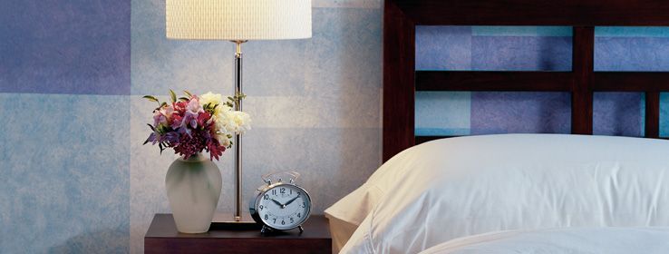 Bedside table with lamp, flowers, and alarm clock next to a bed with white pillows against a textured wall.
