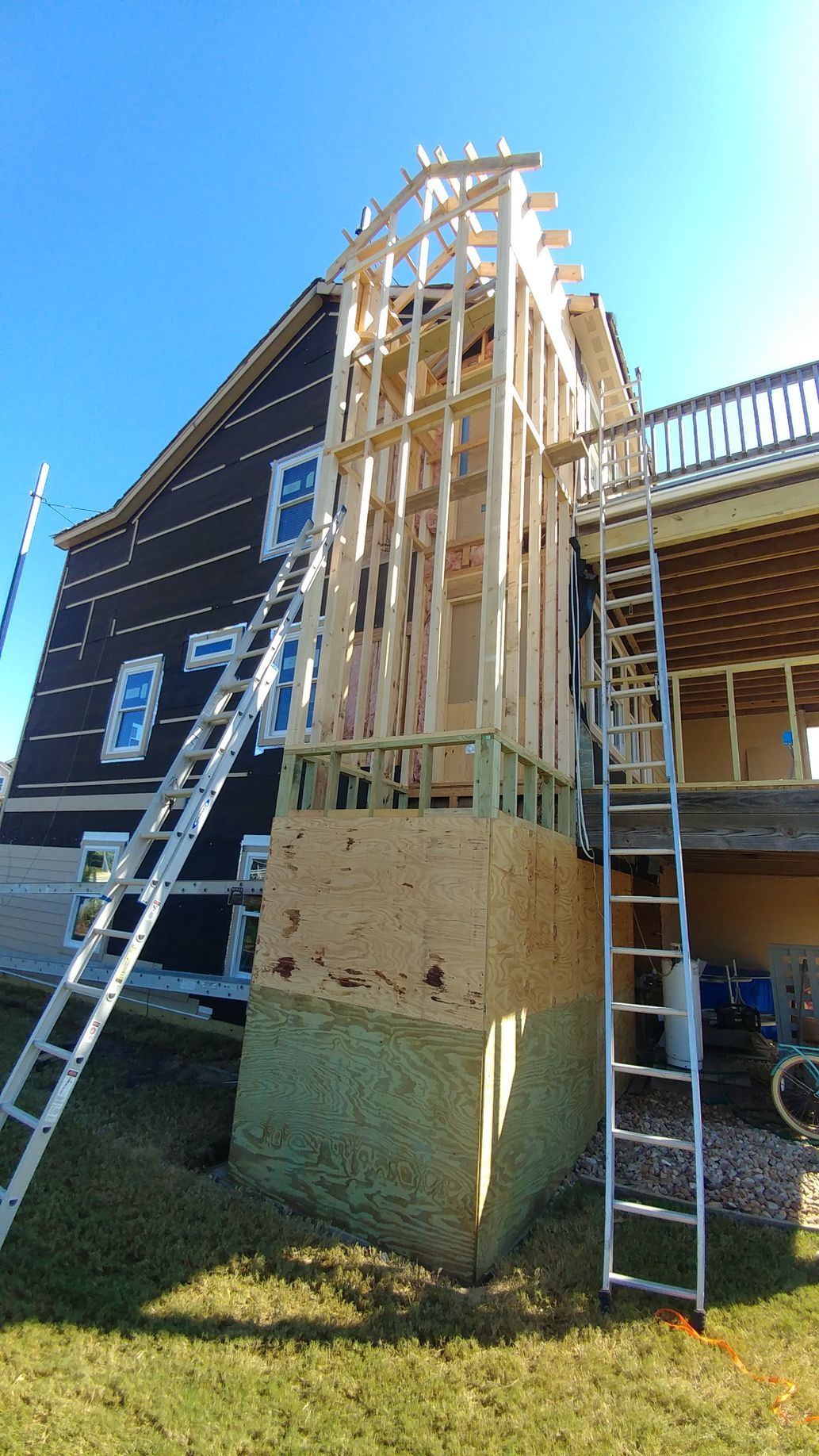Construction site: wooden house addition framework next to a house with brown siding, ladders, and a deck.