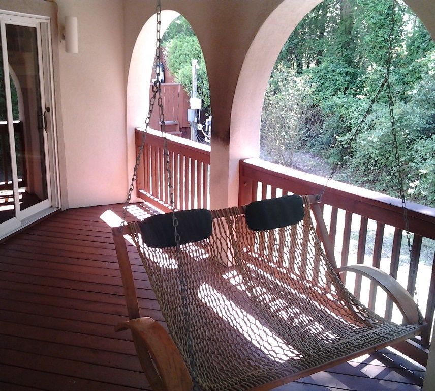 A porch with a hanging hammock, wooden railing, and arched openings leading to a wooded area.