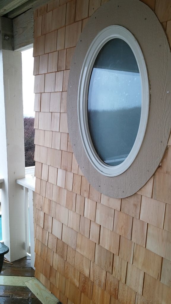 Wall with wooden shingles, an oval window with white trim, and a portion of a white porch.
