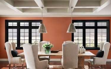 Dining room with orange walls, black-framed windows, a light wood table, and cream-colored chairs.