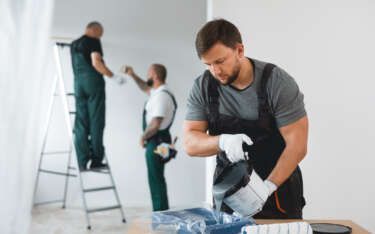Three painters in work clothes painting a white room; one pouring paint.