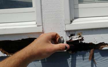 Hand holding a metal object near a badly damaged section of a blue house wall with a window.