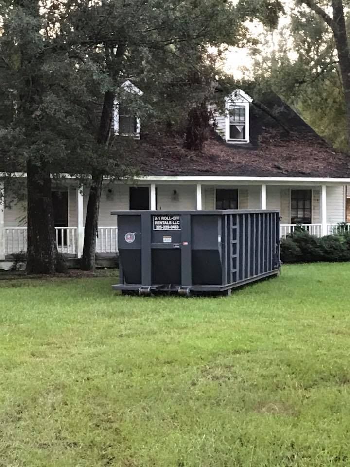 Dark gray dumpster on a lawn in front of a white house with dormer windows.