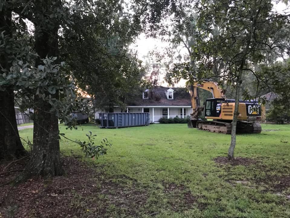 Excavator in front yard of a house, next to a dumpster; green grass, trees.
